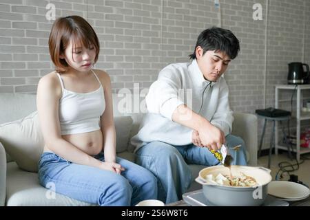 Una donna incinta giapponese sui vent'anni e suo marito, vestito per mostrare i loro stomaci, si siedono su un divano in un appartamento. Foto Stock