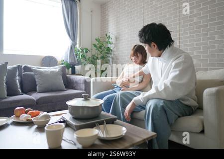 Una donna incinta giapponese sui vent'anni e suo marito, vestito per mostrare i loro stomaci, si siedono su un divano in un appartamento. Foto Stock
