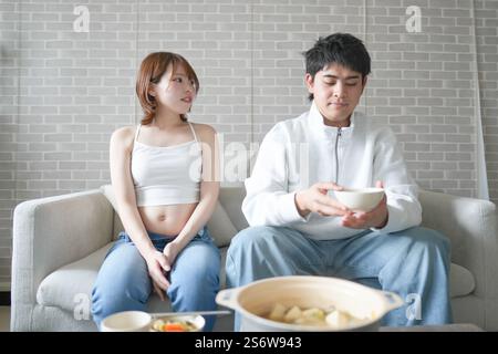 Una donna incinta giapponese sui vent'anni e suo marito, vestito per mostrare i loro stomaci, si siedono su un divano in un appartamento. Foto Stock