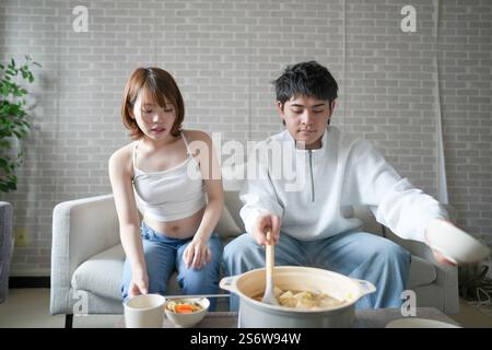 Una donna incinta giapponese sui vent'anni e suo marito, vestito per mostrare i loro stomaci, si siedono su un divano in un appartamento. Foto Stock