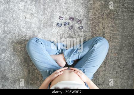 Una donna incinta giapponese di vent'anni, vestita per mostrare il suo stomaco, è seduta su un divano nel suo appartamento, mettendo il palmo sul suo stomaco di fronte Foto Stock