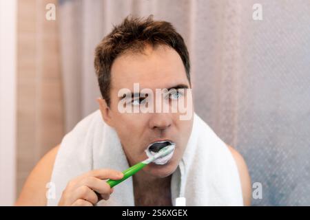 Bel giovane bearded attraente uomo caucasico indossando bianco spugna accappatoio sorride guardando il suo riflesso nello specchio del bagno mentre si spazzolava te Foto Stock
