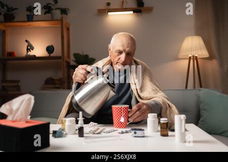 Uomo senatore versare acqua bollente in tazza preparando bevanda calda uomo anziano affetto da febbre virus e freddo in soggiorno di casa Foto Stock