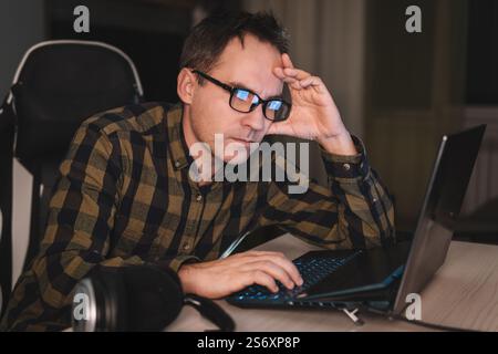 Giovane uomo con una camicia a quadri si è concentrato mentre lavora su un notebook e guarda lo schermo del pc durante il lavoro remoto dall'ufficio domestico, con una tassa di lavoro eccessivo Foto Stock