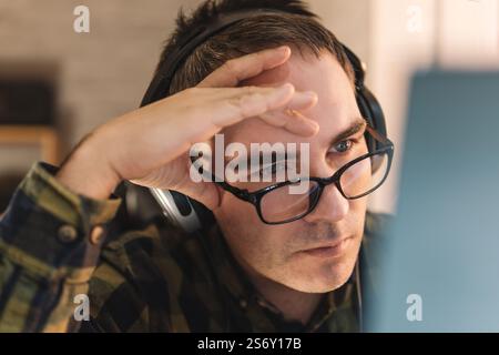 Giovane uomo con una camicia a quadri si è concentrato mentre lavora su un notebook e guarda lo schermo del pc durante il lavoro remoto dall'ufficio domestico, con una tassa di lavoro eccessivo Foto Stock