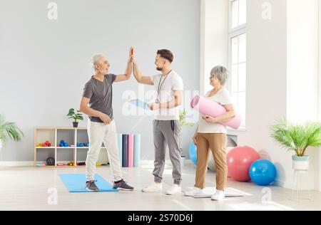 Gli anziani felici e attivi danno un alto cinque al loro istruttore di fitness in palestra Foto Stock