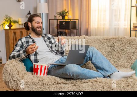 Un giovane che mangia popcorn a casa, saltando occasionalmente i chicchi giocherellando mentre guarda divertenti clip sul laptop. Un ragazzo caucasico si rilassa comodamente sul divano, ridendo ad alta voce di video sciocchi e divertenti Foto Stock