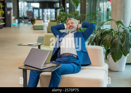 Uomo d'affari felice seduto comodamente con la mano dietro la testa nella moderna hall dell'ufficio chiudendo il computer portatile dopo aver finito il lavoro. Uomo barbuto in tuta che lavora online, e-learning, naviga su Internet su netbook Foto Stock