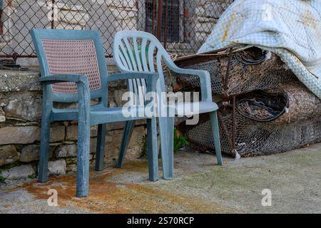 Due sedie in plastica intemprata si trovano accanto a un mucchio di rete metallica arrugginita e a una coperta scartata. La ruggine macchia il terreno. Foto Stock