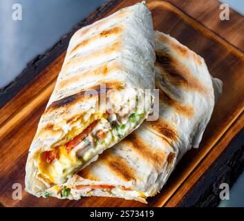 Un burrito appena grigliato ripieno di uova strapazzate, verdure e formaggio è presentato su un piatto di legno. L'esterno grigliato marrone dorato Foto Stock