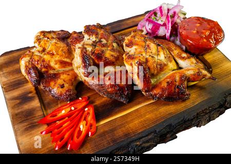 I succosi pezzi di pollo alla griglia sono disposti su un piatto rustico di legno, accompagnati da cipolla rossa fresca, salsa piccante e peperoncino a fette, per creare Foto Stock