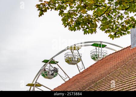 Una ruota panoramica con tre auto su di essa si trova sopra un tetto rosso. Il cielo è nuvoloso e le foglie sull'albero sono verdi Foto Stock
