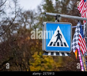 Un segnale di attraversamento pedonale è appeso a un palo. Il cartello è blu e bianco e c'è un uomo che ci cammina sopra. Sono presenti diversi flag appesi al po Foto Stock
