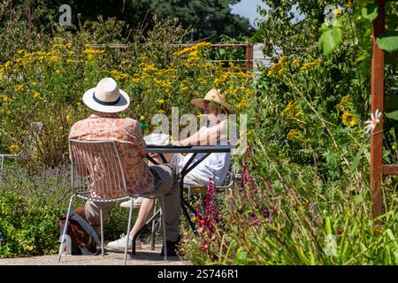 Woking Regno Unito - 13 agosto 2024: Coppia di anziani che indossano cappelli da sole seduti a tavola in giardino circondati da fiori selvatici nelle giornate di sole. Foto Stock
