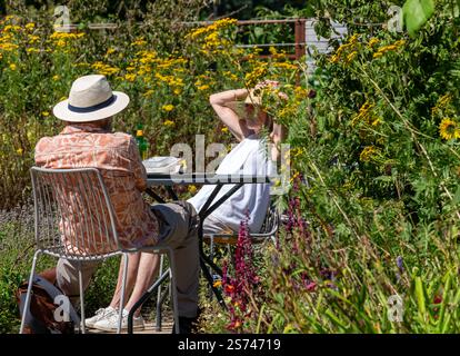 Woking Regno Unito - 13 agosto 2024: Coppia di anziani che indossano cappelli da sole seduti a tavola in giardino circondati da fiori selvatici nelle giornate di sole. Foto Stock