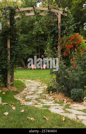 Sentiero in pietra ammiraglia attraverso un manto di legno coperto da Vitis arrampicata - viti che portano a sedie a sdraio rosa Adirondack in giardino rustico sul retro. Foto Stock
