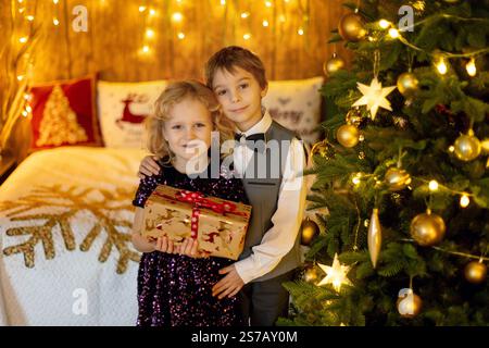 Dolci bambini biondi, fratelli e sorelle, ragazzo e ragazza, vestiti eleganti casual in una casa decorata per Natale, regali ricevuti, felicità Foto Stock
