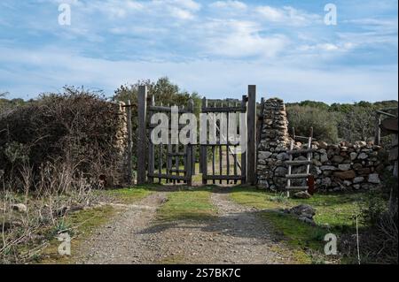 Un cancello in legno rustico con muretti a secco, situato in una campagna rurale con un sentiero sterrato e vegetazione naturale. L'ambiente trasmette tranquillità e. Foto Stock