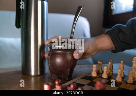 Mano che tiene in mano un tè compagno, durante una partita a scacchi su un comodo divano. Foto Stock