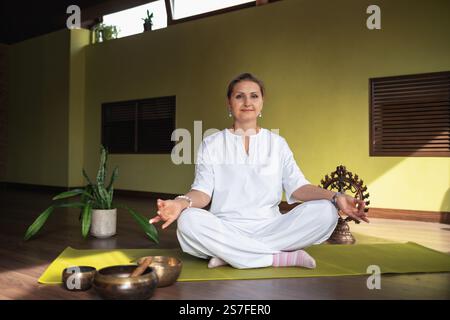Donna che pratica kundalini yoga, meditando seduta in posizione di loto sul tappeto in camera con abiti bianchi, facendo gesti di coscienza con mani e loo Foto Stock
