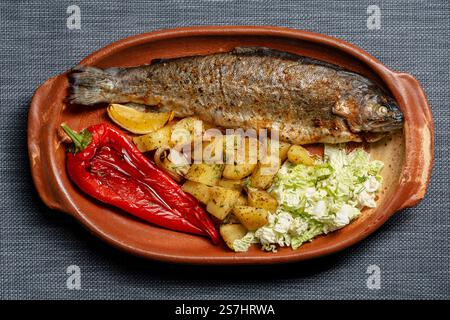 Gustoso pesce di trota al forno con pepe rosso, patate e insalata di cavolo cinese in un piatto di argilla sul tavolo, vista ravvicinata dall'alto Foto Stock
