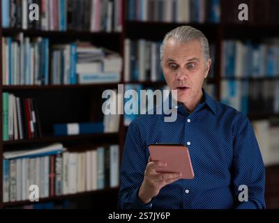 Un uomo che indossa una camicia blu davanti a una libreria reagisce con occhi ampi e sorpresi mentre legge il suo e-book, catturando un momento di discoteca inaspettata Foto Stock