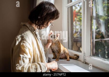 Donna proprietaria di animali domestici che accarezza il gatto devon rex sul davanzale alla luce del sole a casa, godendosi la comunicazione Foto Stock