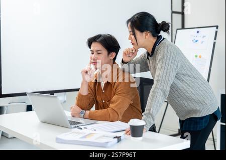 Due uomini d'affari asiatici, concentrati e seri, guardano lo schermo di un notebook, proponendo soluzioni di brainstorming per risolvere i problemi del loro progetto, lavorando insieme io Foto Stock