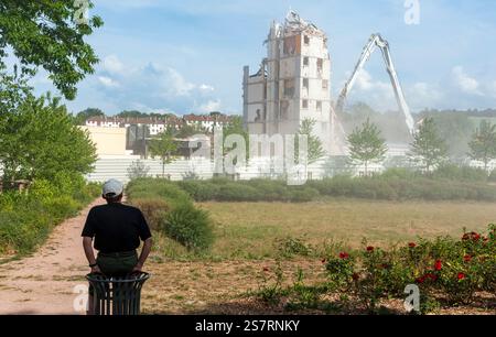Un uomo visto da dietro osserva la demolizione del suo vecchio edificio utilizzando un escavatore dotato di cesoia idraulica montata su un braccio lungo. Foto Stock