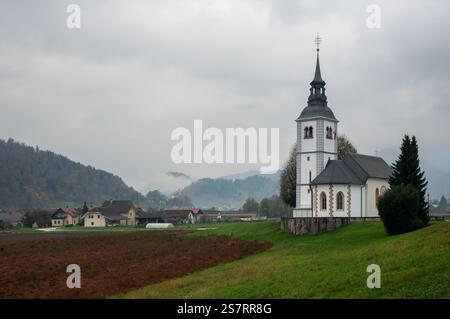 Chiesa bianca con alto campanile in piedi su una collina verde che si affaccia su un piccolo villaggio e campi arati nella campagna slovena. Skofja Loka Foto Stock