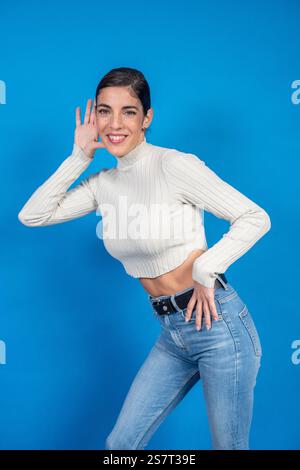 La giovane donna sorride mentre si stringe la mano all'orecchio, ascoltando intensamente su uno sfondo blu vibrante Foto Stock