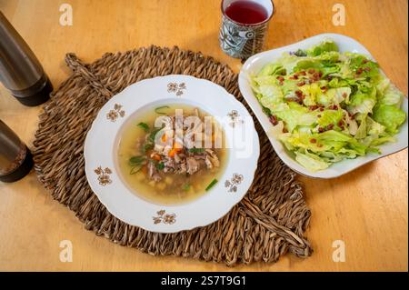 Zuppa di coniglio con verdure nel piatto, insalata di lattuga con pancetta nel recipiente, bamboo pad, tazza di tè e sale e pepe shaker su un tavolo di legno. Foto Stock