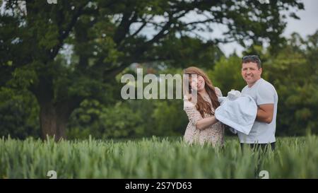 Genitori sorridenti che cullano neonato ondulato, in piedi in un prato lussureggiante, condividendo un momento intimo di legame familiare sotto la luce soffusa del sole Foto Stock