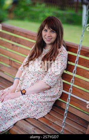 Mamma in attesa riposa su un'altalena in legno, indossa un abito floreale, rilassati nel lussureggiante giardino con un sorriso radioso Foto Stock
