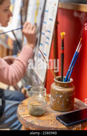I pennelli in un contenitore sono posizionati su uno sgabello e dietro una donna siede a disegnare Foto Stock