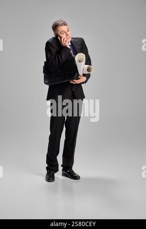 Uomo che parla al telefono tenendo in mano i fogli rotolati e una valigetta, sembra leggermente sopraffatto, bilanciando le attività con la messa a fuoco su sfondo grigio. Foto Stock