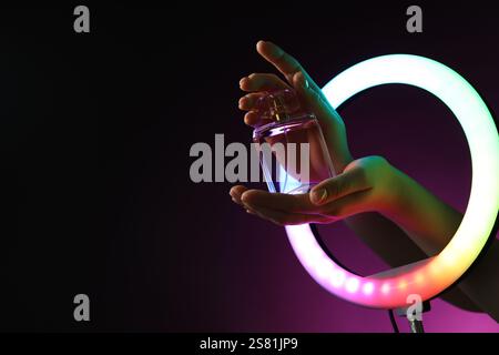 Donna che tiene il profumo attraverso una lampada ad anello su sfondo viola scuro con luci al neon, primo piano. Spazio per il testo Foto Stock