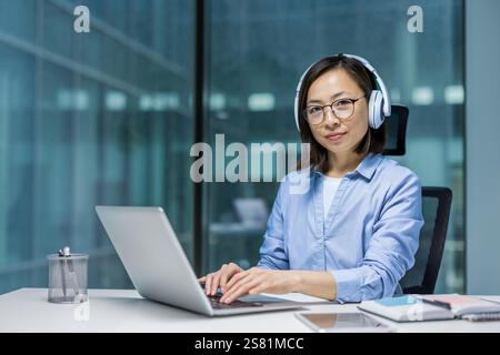 Ritratto di una giovane donna asiatica al suo posto di lavoro all'interno dell'ufficio. La donna è seria e concentrata guardando la telecamera. Lavoratore in ufficio che indossa le cuffie e utilizza un computer portatile. Foto Stock