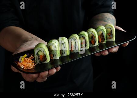 Uno chef presenta un elegante piatto nero con otto sushi all'avocado ordinati ripieni di verdure fresche. I rotoli sono accompagnati da shr Foto Stock