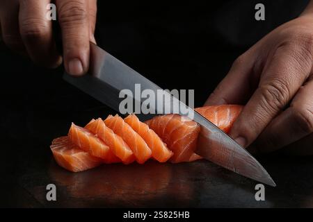 Uno chef affetta filetto di salmone fresco con un coltello giapponese affilato, mostrando precisione e tecnica su uno sfondo scuro. Foto Stock