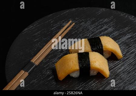 Tamago Nigiri presenta dolci fette di omelette giapponesi poste sopra il riso sushi e legate con una striscia di nori, presentate elegantemente su un piatto scuro con ch Foto Stock