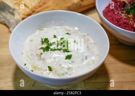 Salse salutari a base di yogurt vegano con cetriolo, aglio ed erbe aromatiche con guarnigione di prezzemolo in una ciotola bianca con pane su un rustico asse di legno, fatto in casa Foto Stock