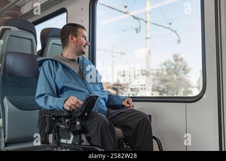 Viaggiatore di sesso maschile con disabilità che si gode la vacanza e il viaggio in treno in Europa, seduto su una sedia a rotelle che guarda fuori dal finestrino. Foto Stock