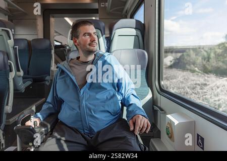 Viaggiatore di sesso maschile con disabilità che si gode la vacanza e il viaggio in treno in Europa, seduto su una sedia a rotelle che guarda fuori dal finestrino. Foto Stock