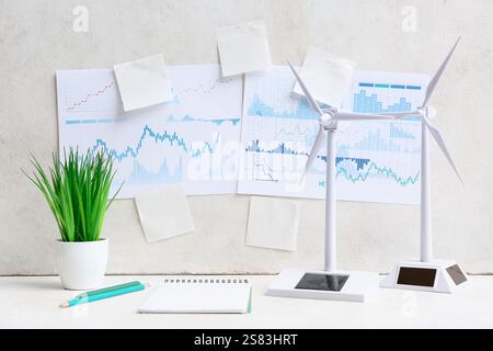 Diagrammi con note adesive vuote e modelli di turbine eoliche sul tavolo vicino alla parete illuminata. Concetto di energia verde Foto Stock