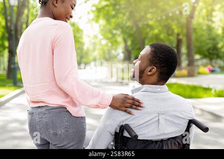 Giovane donna nera e il suo ragazzo disabile in sedia a rotelle trascorrere il tempo insieme all'aperto, vista posteriore Foto Stock