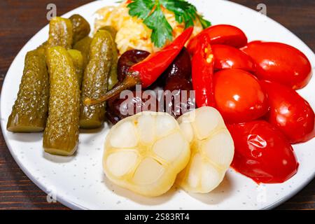 Una colorata disposizione di verdure sottaceto, tra cui cetriolini, peperoni e pomodori, è esposta su un piatto bianco. Le erbe fresche si aggiungono al vibrante p Foto Stock