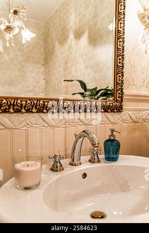 Elegante lavandino del bagno con lavabo in ceramica bianca, rubinetto in argento con sfumature dorate, specchio con cornice dorata, piastrelle con motivi beige ed elementi decorativi raffinati. Foto Stock