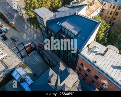 Vista aerea di un edificio moderno con tetto in metallo scuro e finestre in vetro, circondato da vecchie strutture in mattoni, una strada stretta e auto parcheggiate. Foto Stock