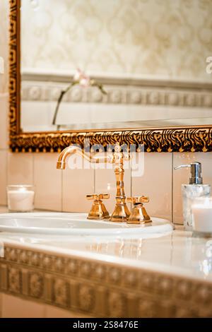 L'area del lavandino del bagno presenta un rubinetto dorato, un intricato specchio incorniciato in oro, spruzzi di piastrelle a motivi geometrici, orchidee e candele accese per un'atmosfera calda. Foto Stock